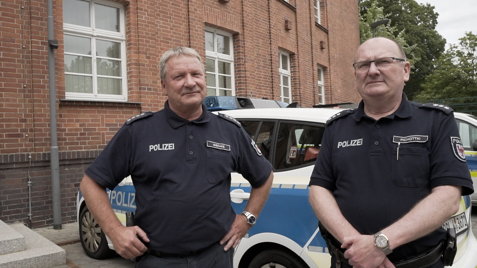 Bild: Teamfoto von Herrn Mischke und Herrn Pichottki vor dem Polizeigebäude - im Hintergrund ein Polizeifahrzeug