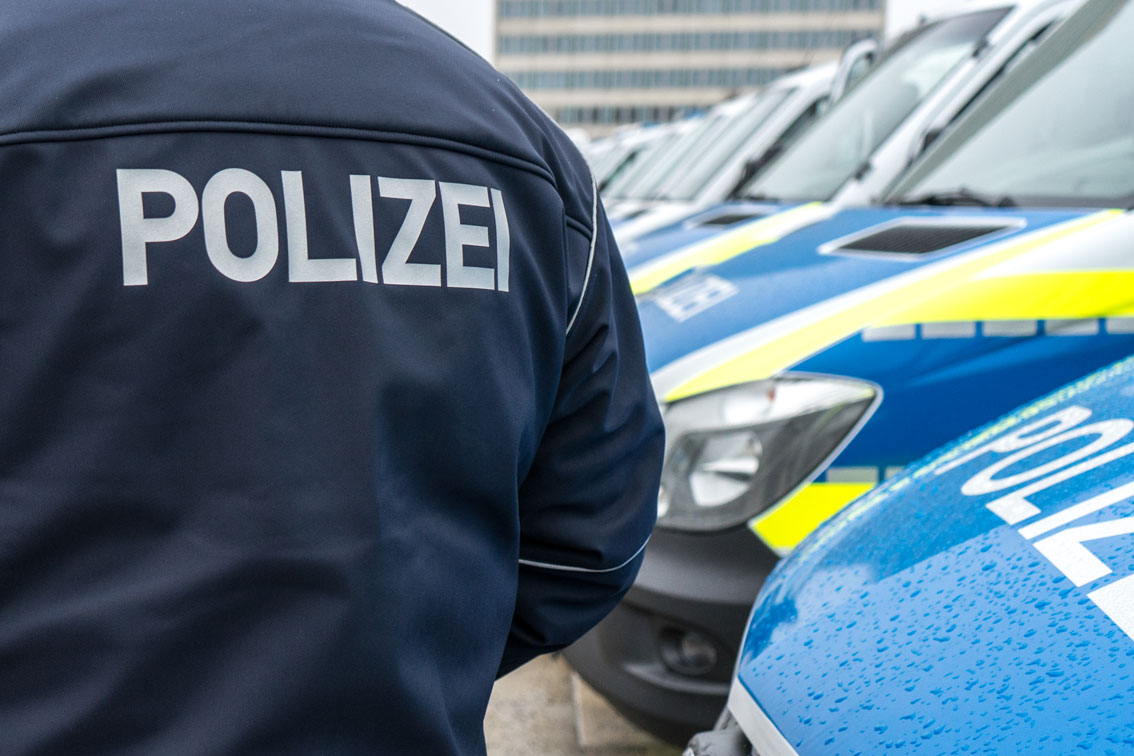 Teaser Polizei Fahrzeuge Rücken eines Polizeibeamten mit der Aufschrift Polizei und Funkstreifenwagen in Hintergrund