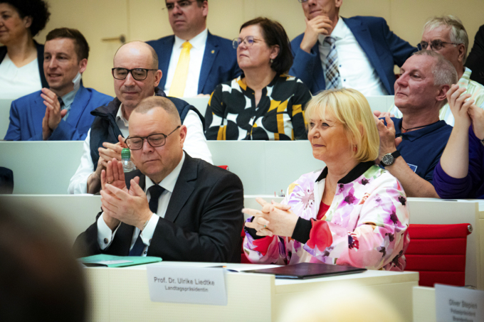 Foto von Landtagspräsidentin Prof. Dr. Liedtke und Minister Stübgen auf ihren Plätzen im Plenarsaal mit Gästen im Hintergrund sitzend